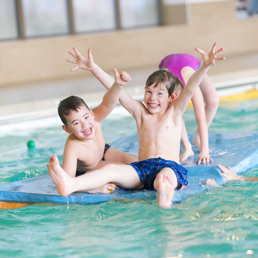 Een groepje blije jongens en meiden balanceren op een drijfmat in het water van het zwembad