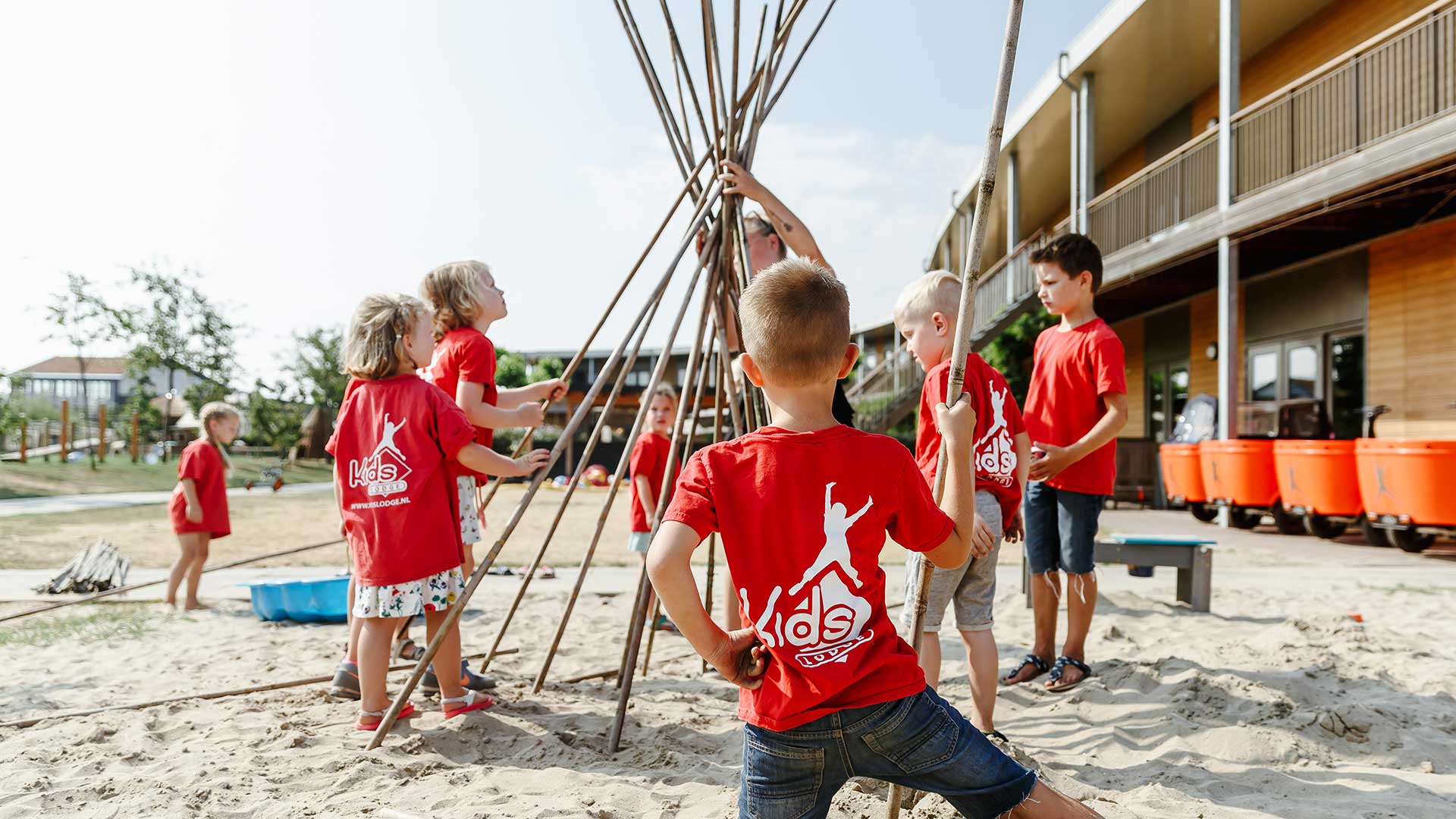 Kinderen dragen Kids Lodge t-shirts en bouwen een tipi van lange stokken in de zandbak van locatie Jeugdland in Nieuwegein